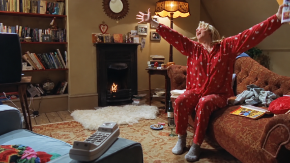 A woman wearing red pajamas with arms wide open sits on a couch in a living room with a fireplace.