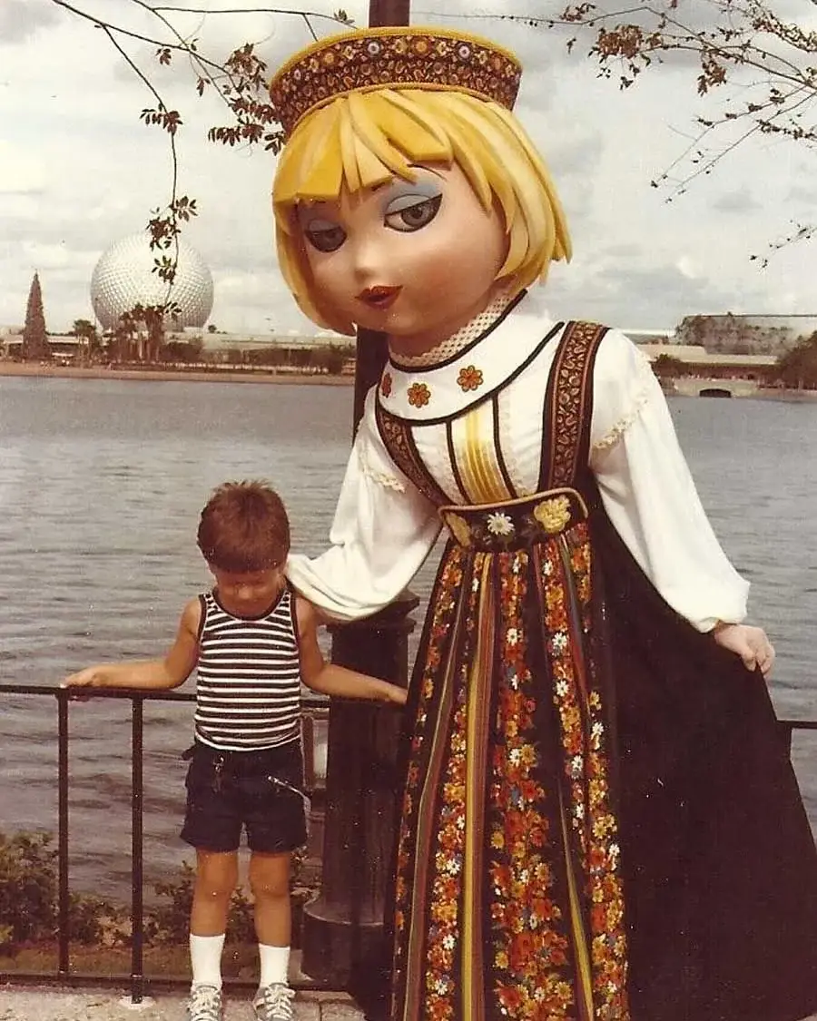 A child in a striped shirt stands next to a large blonde doll mascot in traditional dress by a lakeside, with domed buildings visible in the background.