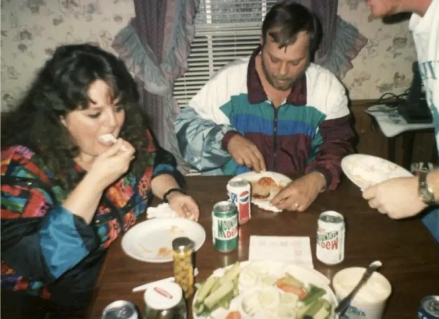 Two people sit at a table eating, with soda cans, veggie platter, and dip. One person feeds themselves with a fork, while another cuts food. A third person is partially visible, serving food.
