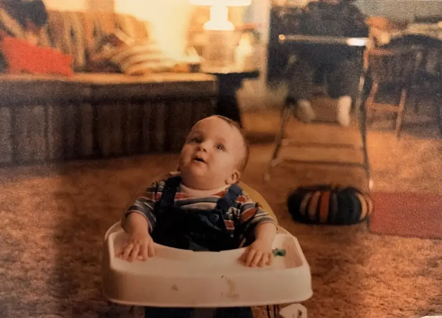A baby in a walker looks up, wearing striped clothes, on a brown carpet in a cozy, warmly lit living room with a couch and another person in the background.