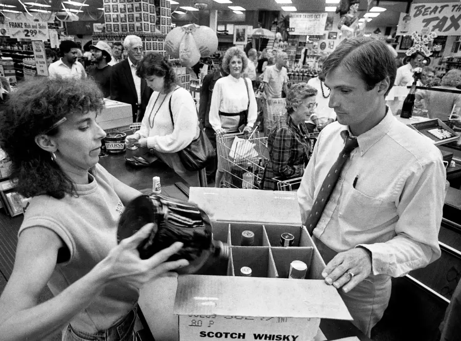 A woman places a large bottle of liquor into a box labeled "Scotch Whisky" at a busy store counter while a man in a tie watches. Several people wait in line and shelves are stocked in the background. Black and white photo.