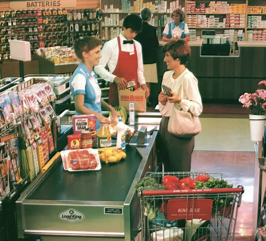 A woman smiles while checking out groceries at a supermarket. A cashier and bagger assist her. The conveyor has meat, cereal, bananas, and juice. The cart contains produce. Shelves and products are visible in the background.