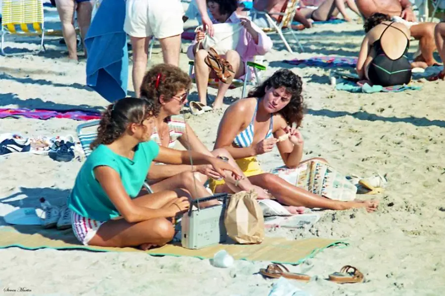 Three women sit on a beach towel, talking and eating snacks, surrounded by beachgoers. Sandals, bags, and other towels are scattered around them on the sandy beach.