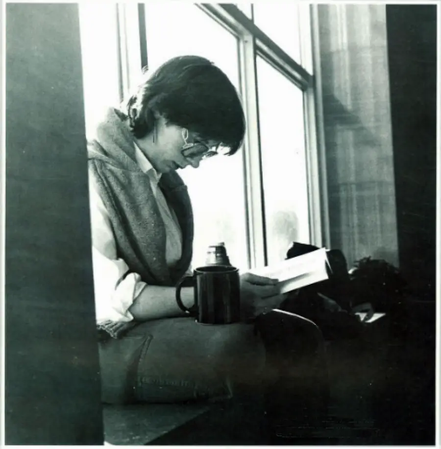 A person with glasses sits by a large window, reading a book. They wear a sweater draped over their shoulders and have a thermos placed in front of them. Light streams in through the window, creating a calm atmosphere.