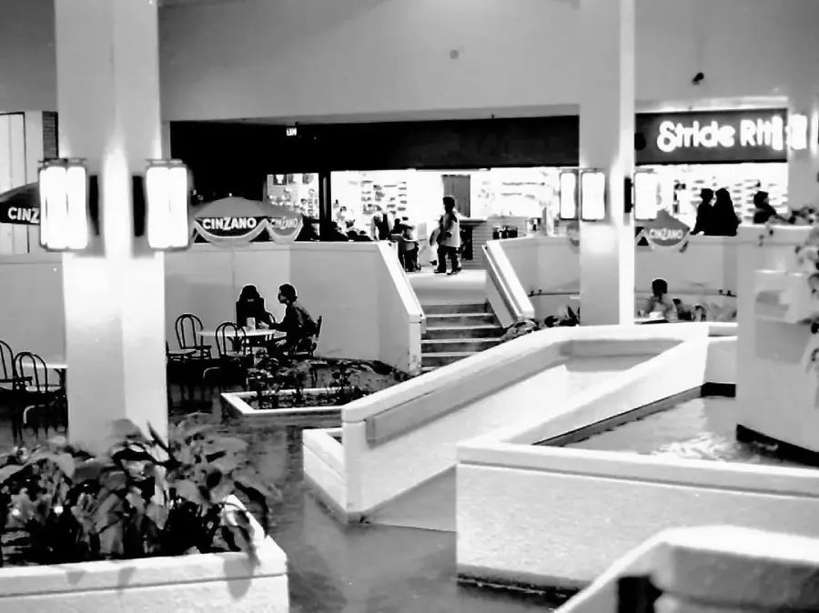 Black-and-white photo of a mall interior with people sitting at tables near a small indoor water feature. Stores, including Stride Rite, are visible in the background. Plants and stairs are also present in the scene.
