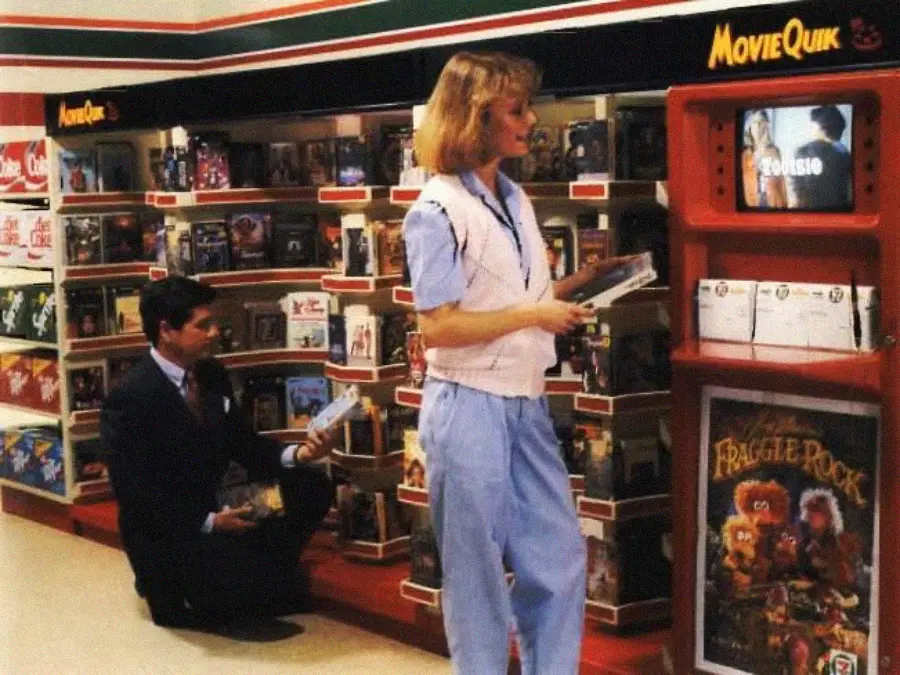A woman and a man browse VHS tapes at a 1980s video rental section in a store. The woman stands holding tapes, while the man kneels. A small TV screen displays a video preview above shelves of movies.