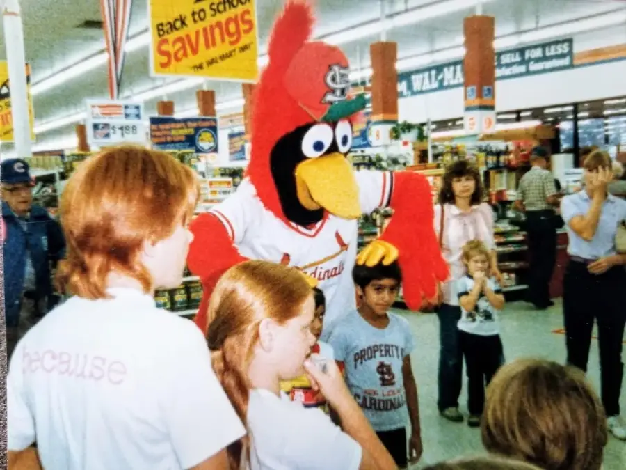 A person in a red Cardinals mascot costume stands in a store surrounded by children and adults, with “Back to School savings” signs hanging above the aisles.