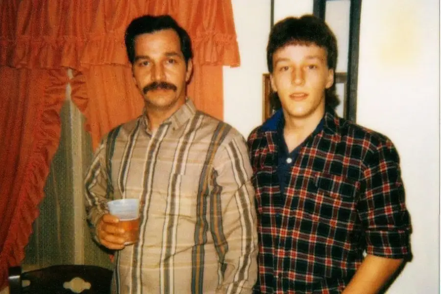 Two men stand side by side indoors; one has a mustache, holds a drink, and wears a striped shirt, while the other wears a plaid shirt. Red curtains and a wall are visible in the background.
