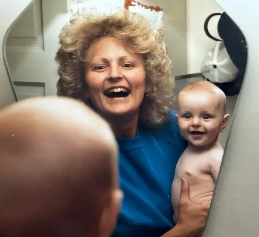 A smiling woman holding a happy, shirtless baby stands in front of a mirror. Both their faces are reflected clearly, while the back of the baby's head is seen in the foreground.