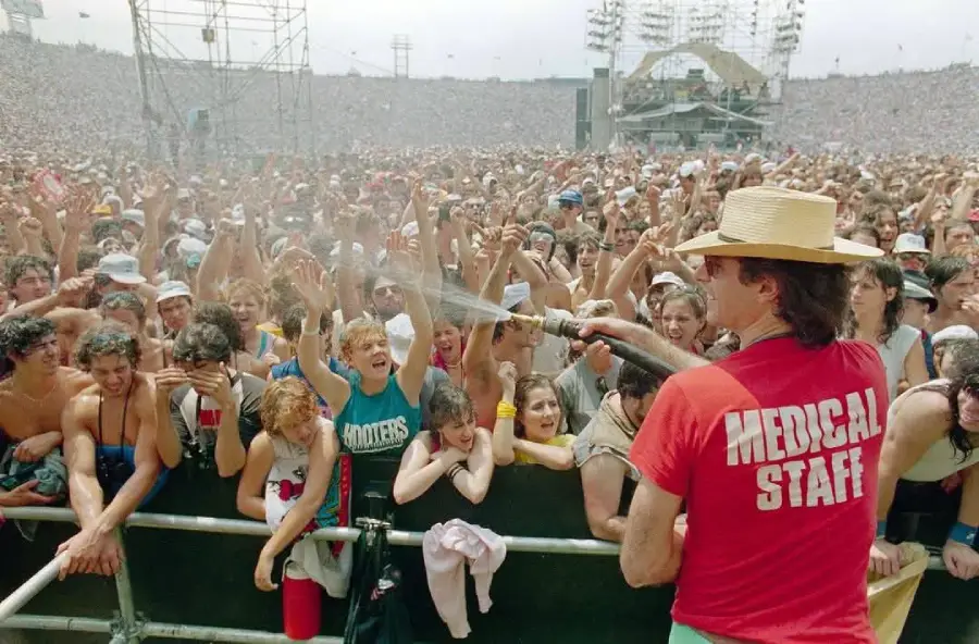 A man in a "Medical Staff" shirt sprays water over a large, excited crowd at an outdoor concert on a sunny day. The audience smiles and cheers, many reaching out toward the spray.