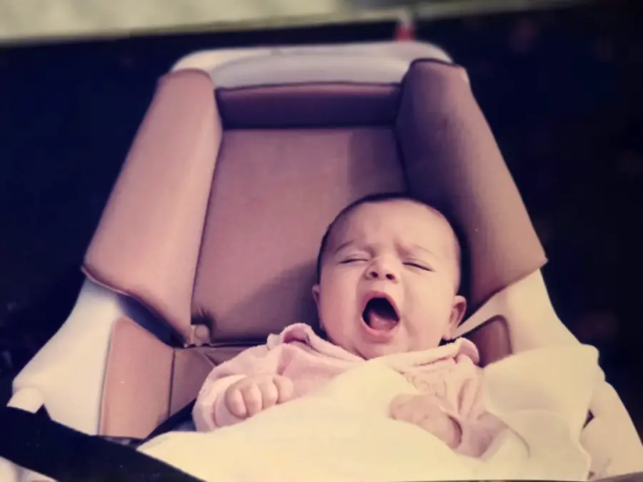 A baby in a car seat yawning, dressed in a light pink outfit and covered with a white blanket.