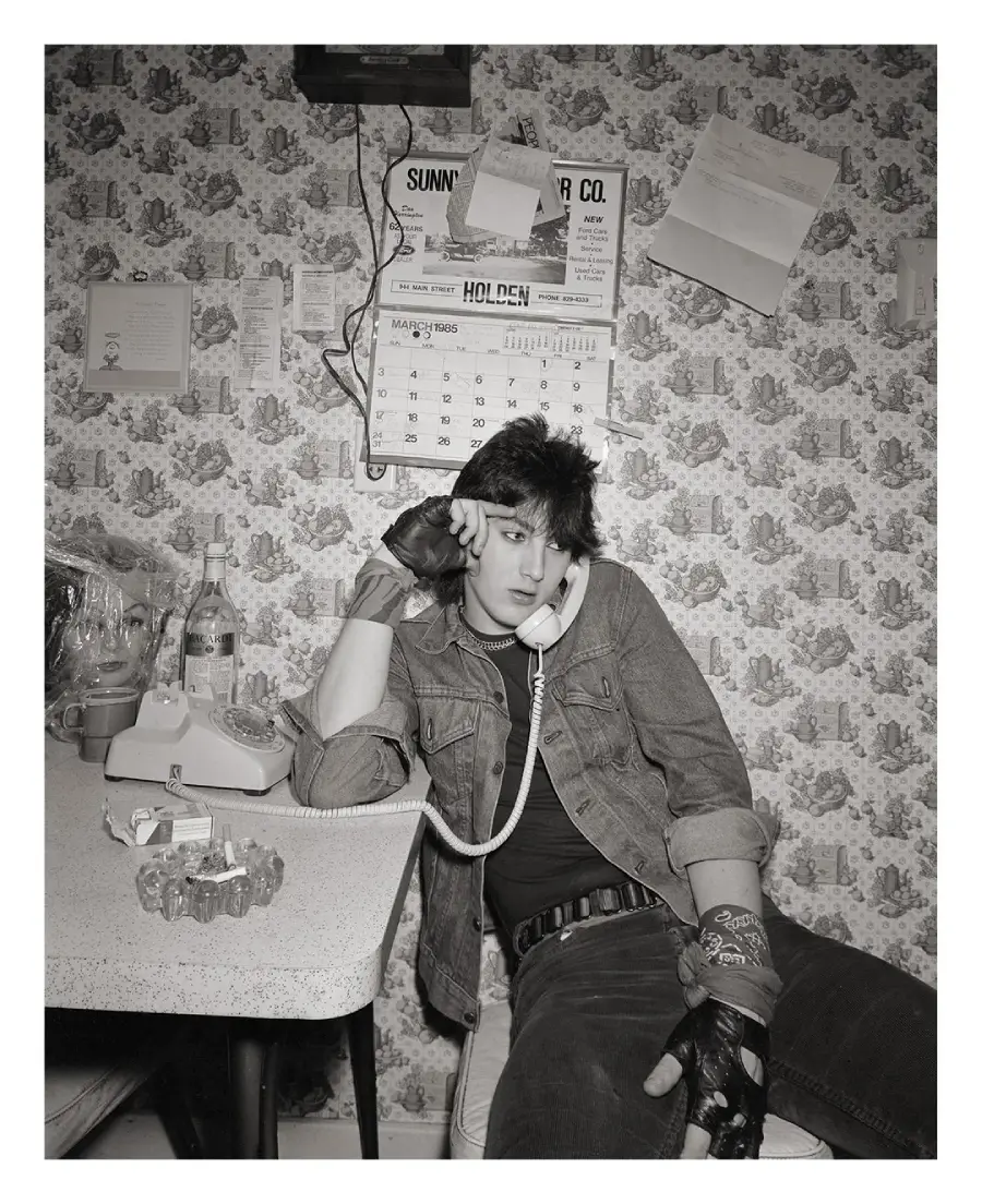 A person in a denim jacket and gloves sits by a table, talking on a corded phone. The background features patterned wallpaper, a calendar, and papers pinned to the wall.