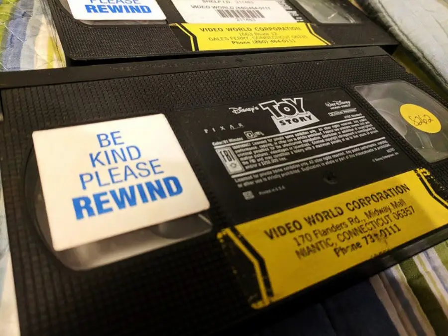 Close-up of Toy Story VHS tape with a sticker that says “BE KIND PLEASE REWIND.” Another VHS tape is partially visible beneath it. Both tapes rest on a striped surface.