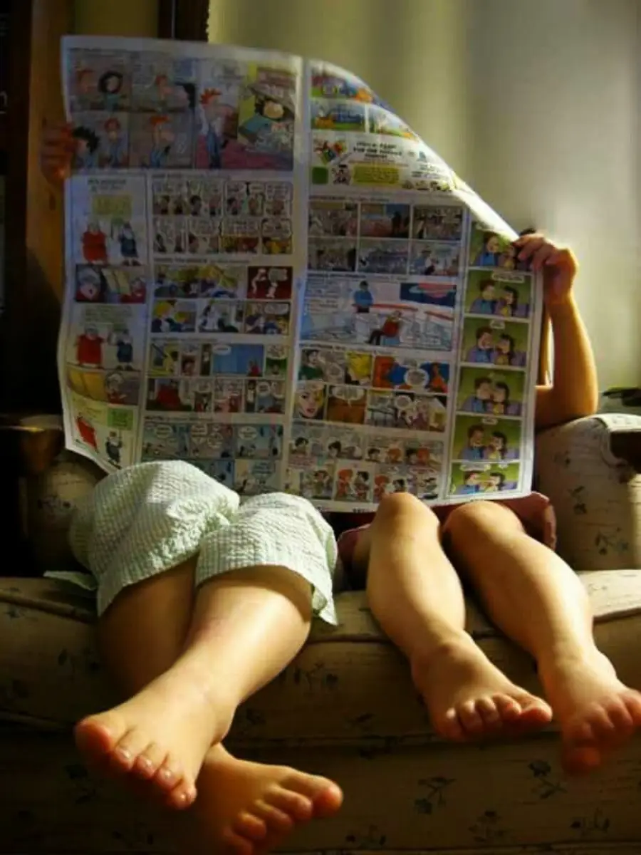 Two children sit on a couch with their bare feet propped up, reading a large newspaper comic section that covers both of their faces. Sunlight softly illuminates the scene.
