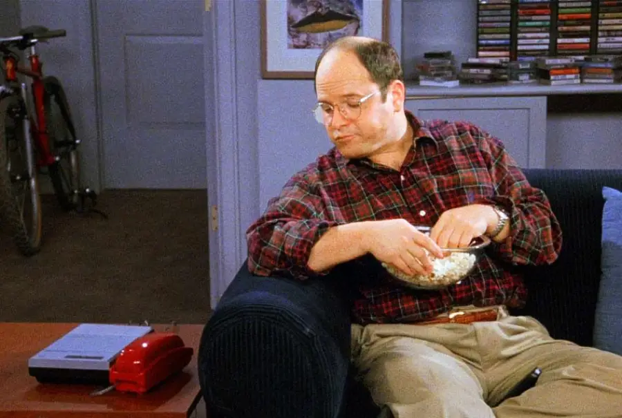 A man in glasses and a plaid shirt sits on a couch eating popcorn, looking at a red phone on a table. A bicycle is visible in the background.