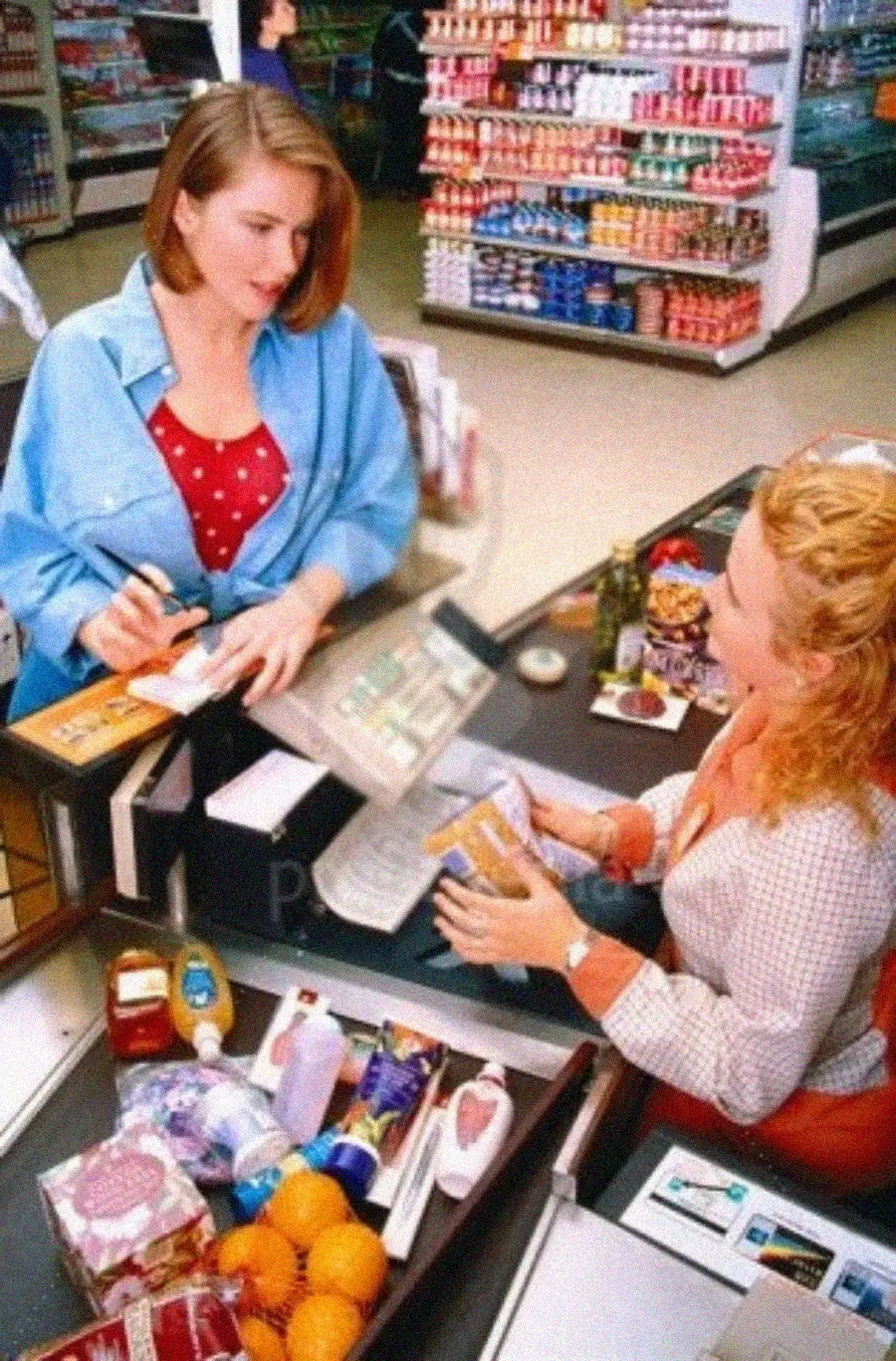A woman in a blue shirt stands at a grocery store checkout counter while another woman scans groceries. Items like oranges, milk, and snacks are on the conveyor belt. Shelves with products fill the background.