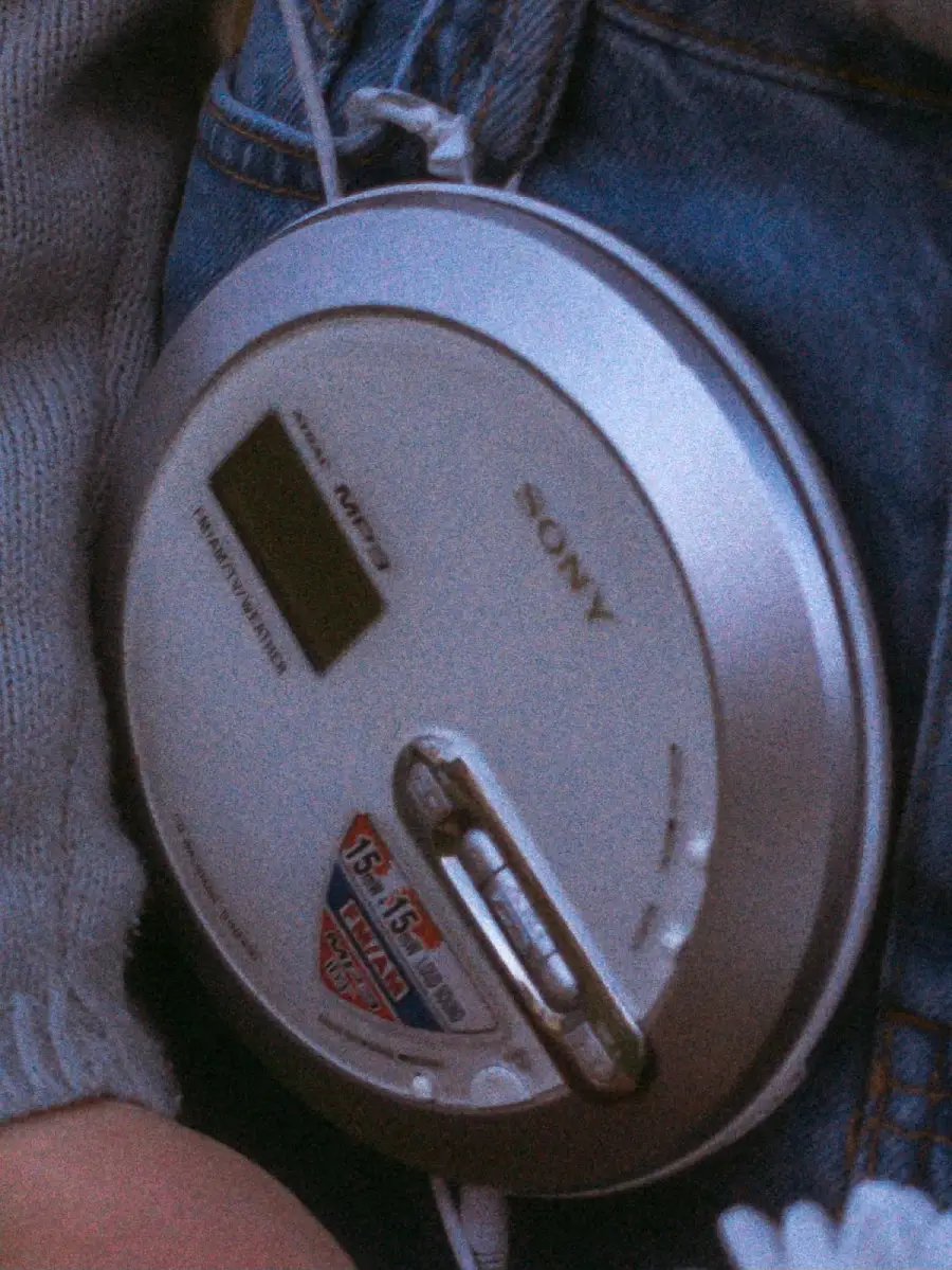 A close-up of a silver Sony portable CD player attached to a person's jeans, with part of a sweater sleeve visible. Earphone wires are plugged into the player.