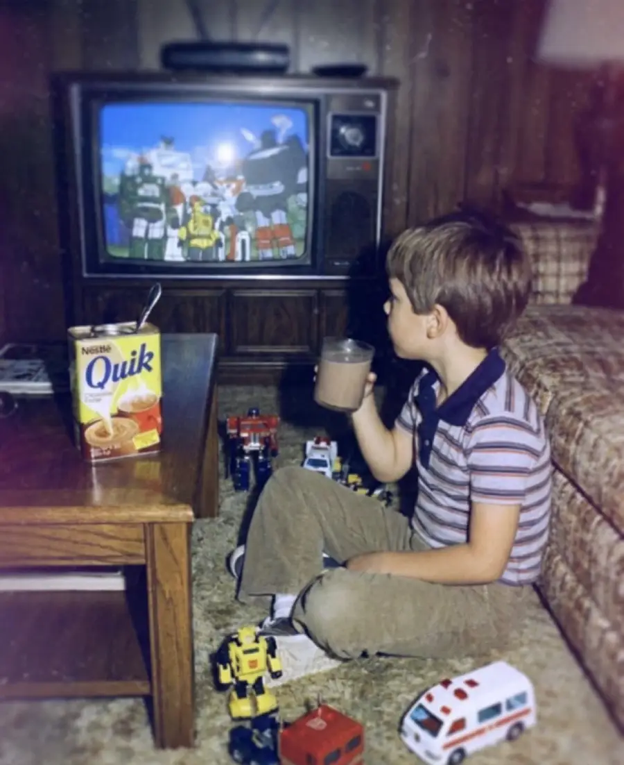 A young boy drinks chocolate milk while watching Transformers on a CRT TV. Transformers toys and a box of Nestle Quik are on the table and floor. The setting appears retro, with wood-paneled walls and vintage furniture.