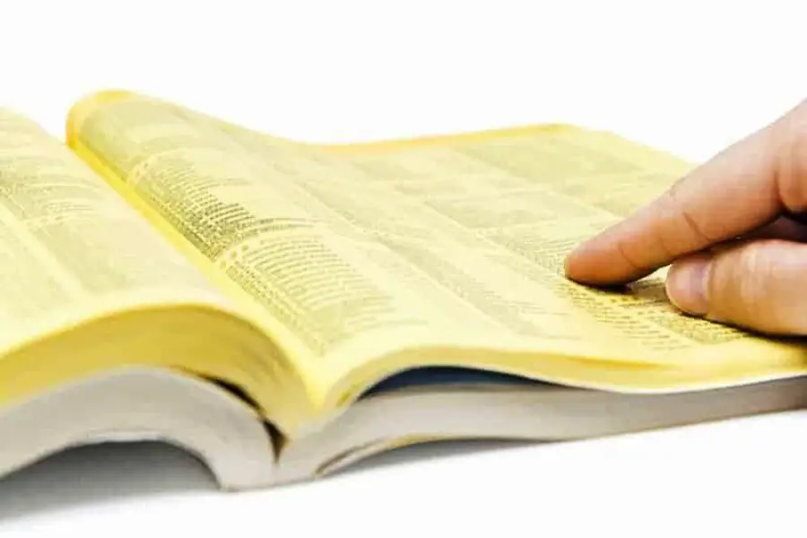 A hand points to a listing in an open yellow pages phone book, with text columns visible on the yellow paper.