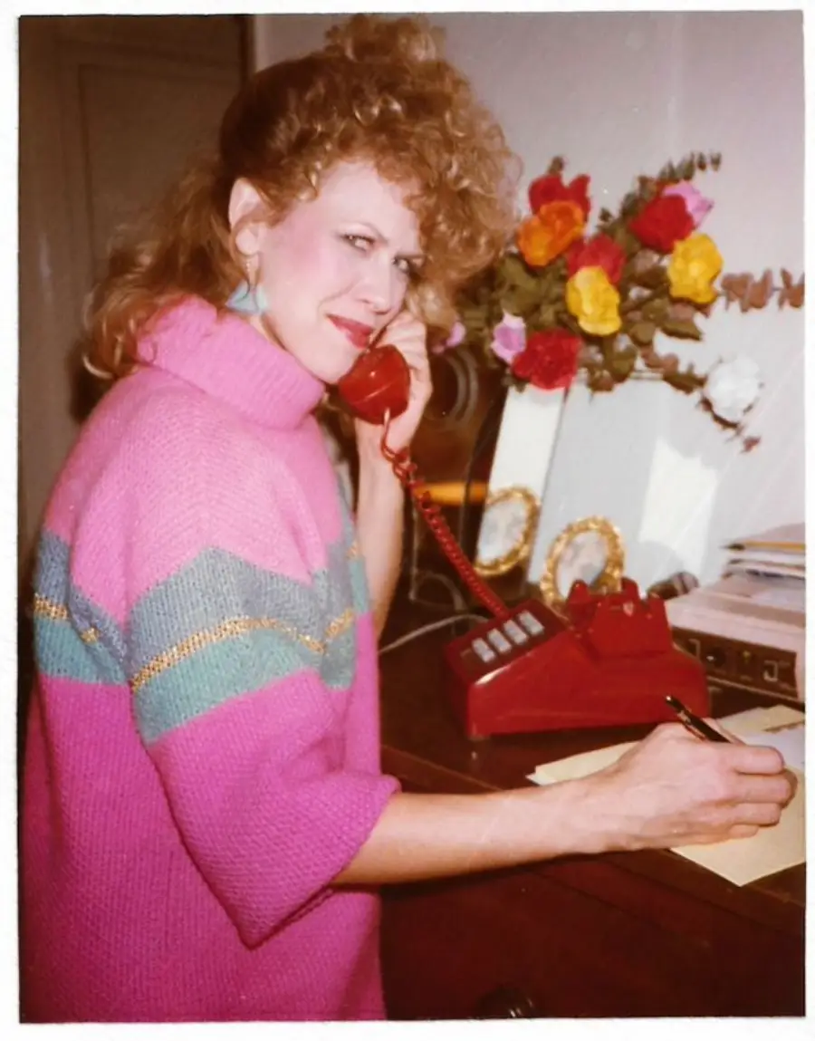Woman in a pink sweater smiles while talking on a red corded phone and writing on paper. There are flowers and framed photos on the wooden desk. The scene appears to be from the 1980s.