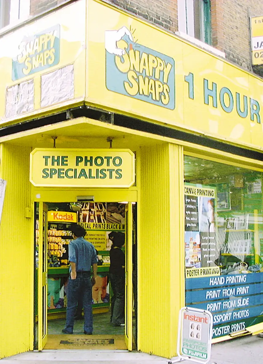 A bright yellow "Snappy Snaps" photo shop with "1 Hour" and "The Photo Specialists" signs. Two people stand inside near photo developing displays and Kodak film.