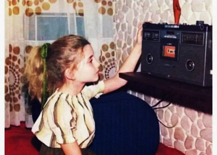 A young girl with a green hair ribbon reaches to press play on a cassette tape player in a retro-styled room with patterned curtains and wallpaper.