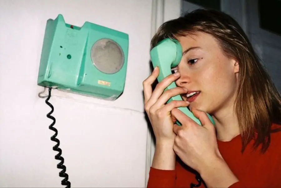 A woman in a red shirt holds a mint-green wall phone with the receiver pressed to her eye instead of her ear, looking curious or playful.