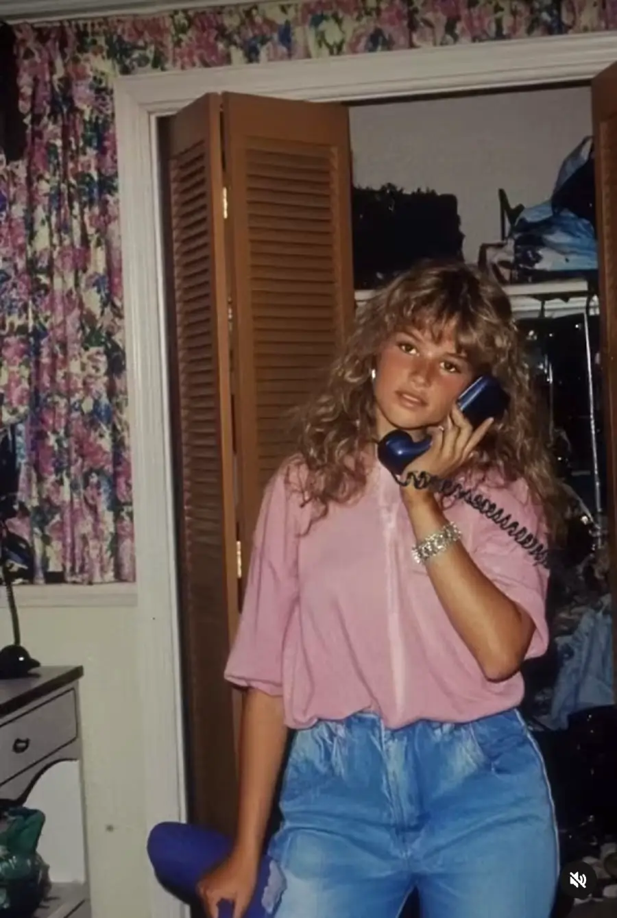 Teen girl with wavy hair, wearing a pink top and blue jeans, talks on a corded phone. She stands in a bedroom with floral curtains and an open closet door in the background.