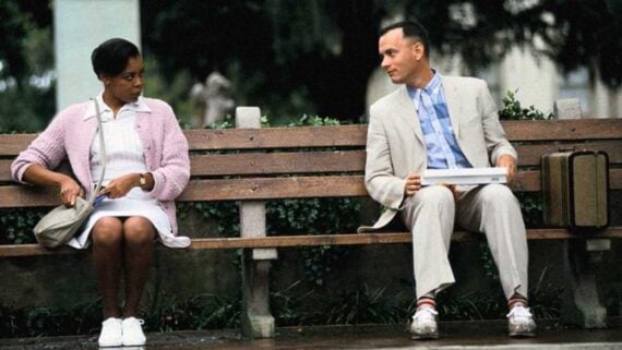 Tom Hanks as Forrest Gump sitting on the famous park bench holding a suitcase.