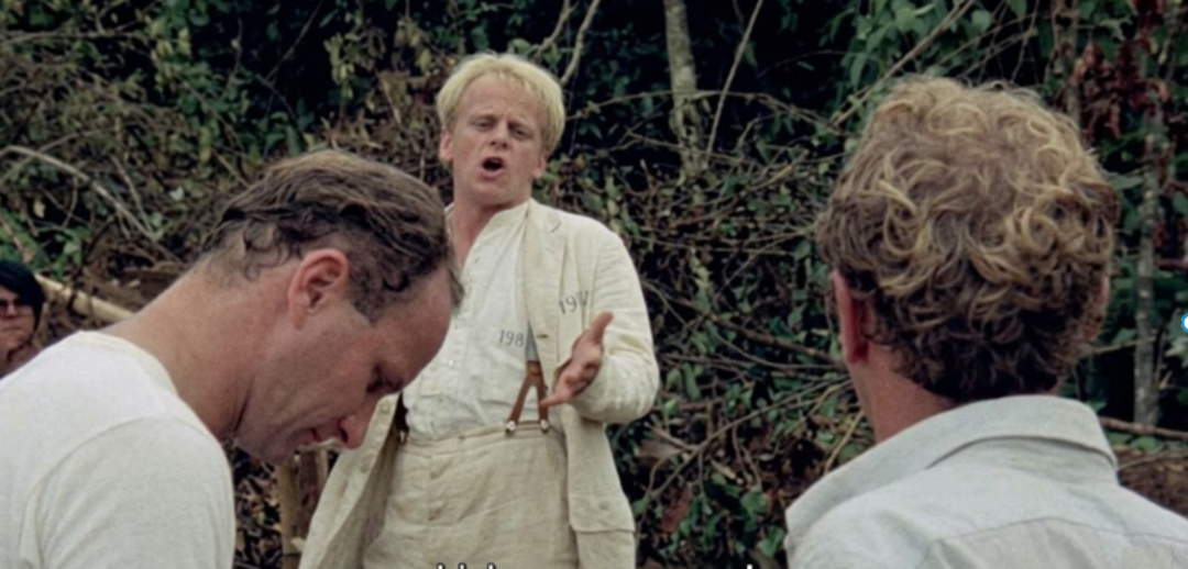 Three men stand outdoors near dense foliage. The man in the center, wearing a white suit with suspenders, gestures expressively with one hand while speaking. The two other men are shown from behind in the foreground.