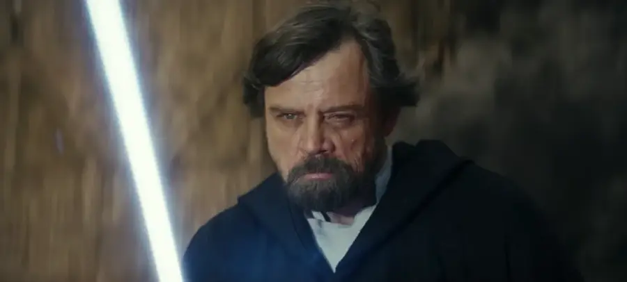 A bearded man in a dark hooded robe holds a glowing lightsaber, looking intensely forward. The background is blurred with brown and gray tones, suggesting action or tension.