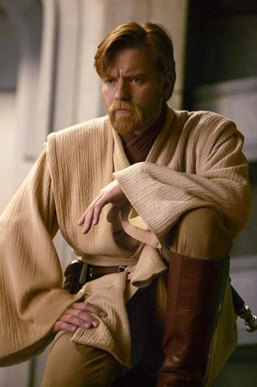 A person in tan Jedi robes with a belt and boots sits on a step, looking serious, with short light brown hair and a beard, in a scene resembling a Star Wars setting.