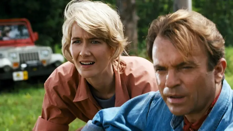 Two people look intently at something off-screen outdoors, with a red jeep in the background and greenery around them. Both have concerned expressions and are dressed in casual clothes.