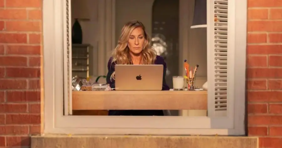 A woman with long blonde hair sits at a desk by an open window, working on a laptop. The desk has pens, papers, and a glass of milk. Brick walls frame the window.
