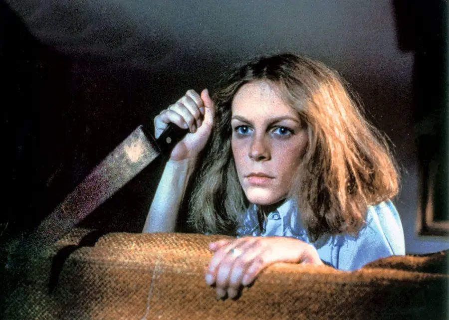 A woman with shoulder-length hair holds a large knife while crouching behind a couch, looking tense and determined in a dimly lit room.