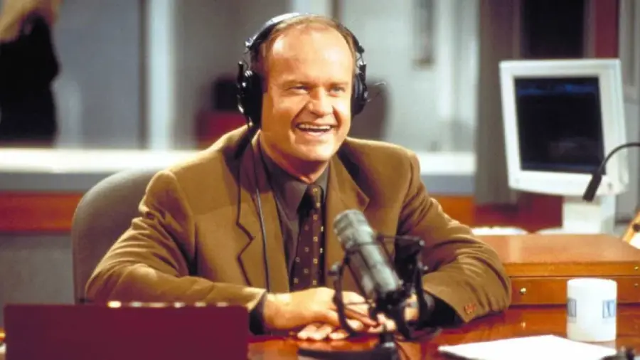 A man in a brown suit sits at a radio studio desk, wearing headphones and smiling while speaking into a microphone. A computer monitor and mug are visible in the background.