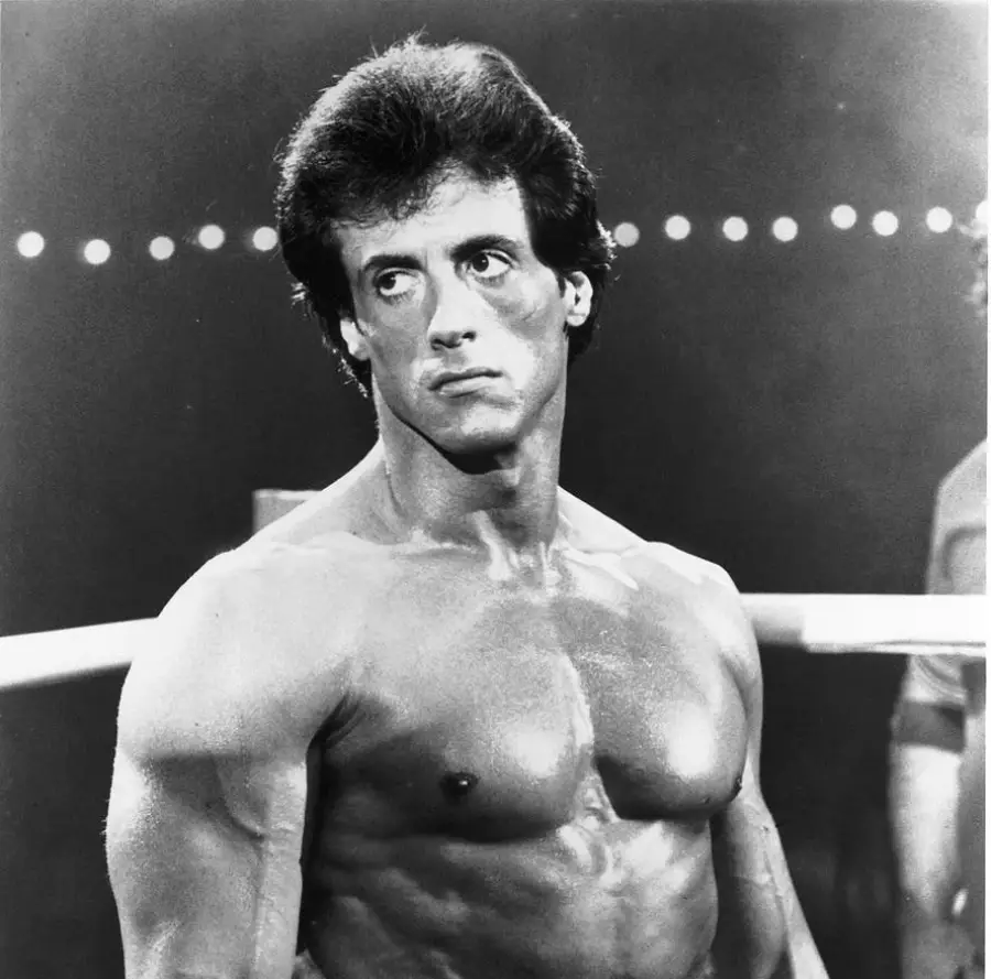 A muscular man stands shirtless in a boxing ring, looking serious. The background is dark with a row of lights behind him.