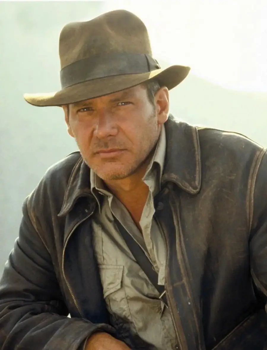 A man in a fedora hat and leather jacket stares ahead with a serious expression. He is wearing a collared shirt and is shown from the chest up against a light background.
