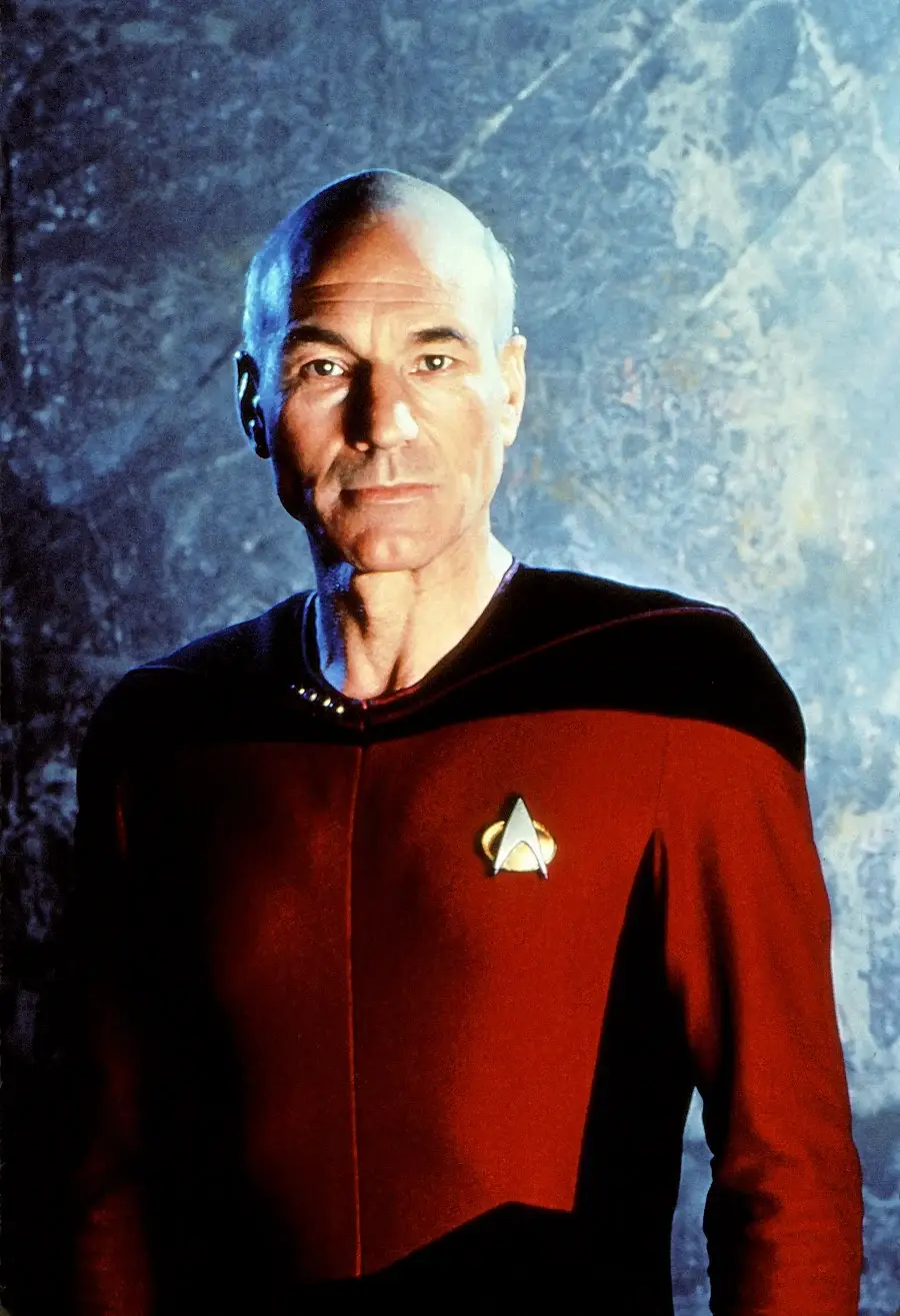 A person in a red and black Starfleet uniform stands against a textured blue-gray background, wearing a gold insignia badge on their chest.