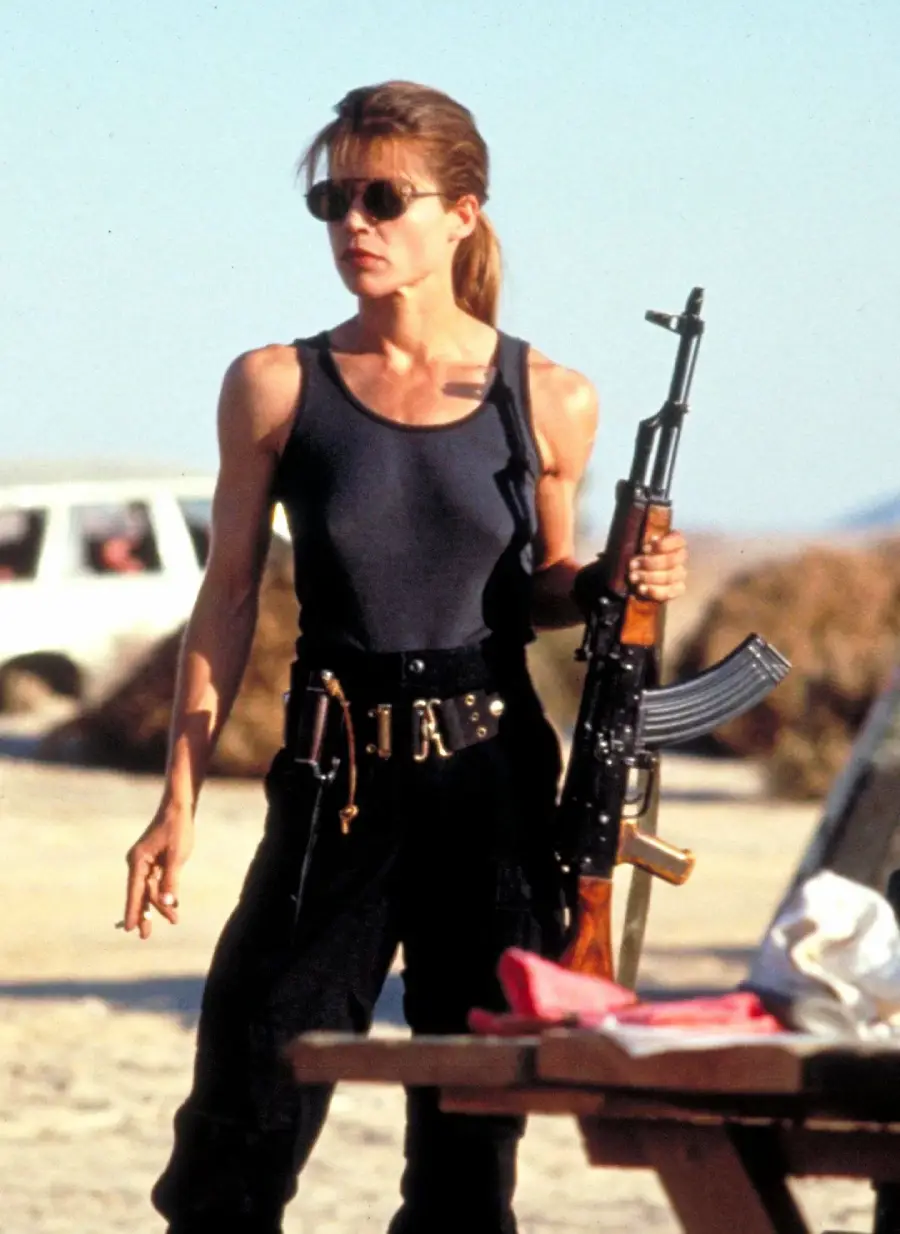 Woman in sunglasses and a black tank top holding a rifle stands outdoors near a table, with a car and dry landscape in the background.