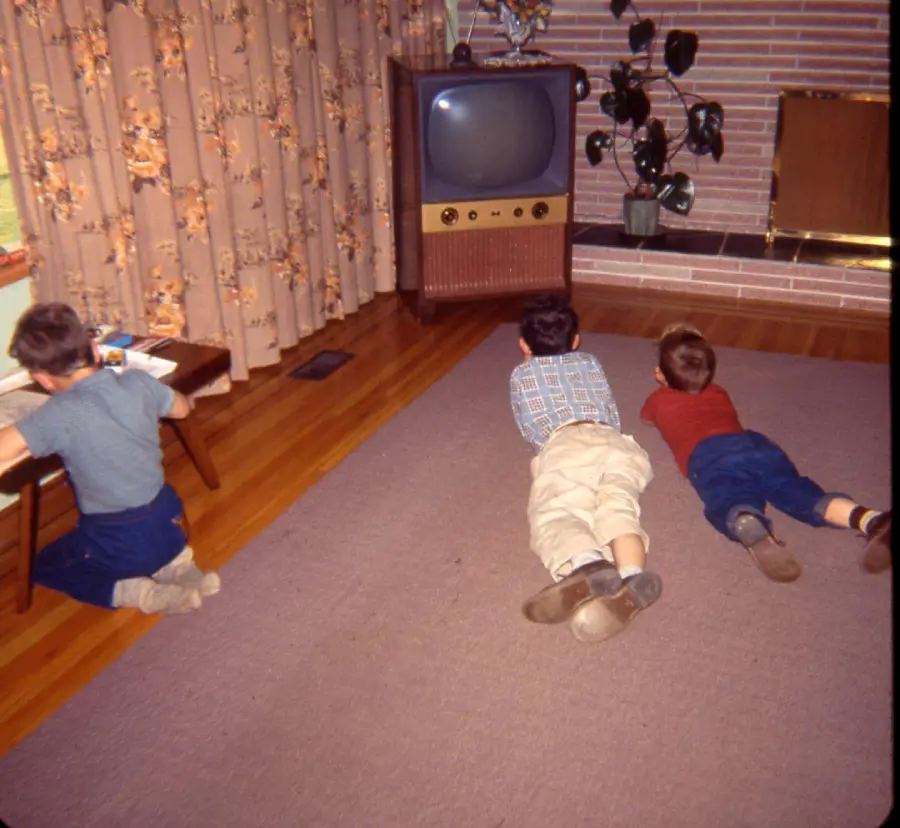 Three children in 1960s-style clothes, two lying on a carpeted floor watching an old TV, while one sits at a small table drawing. The room features floral curtains and a brick fireplace.