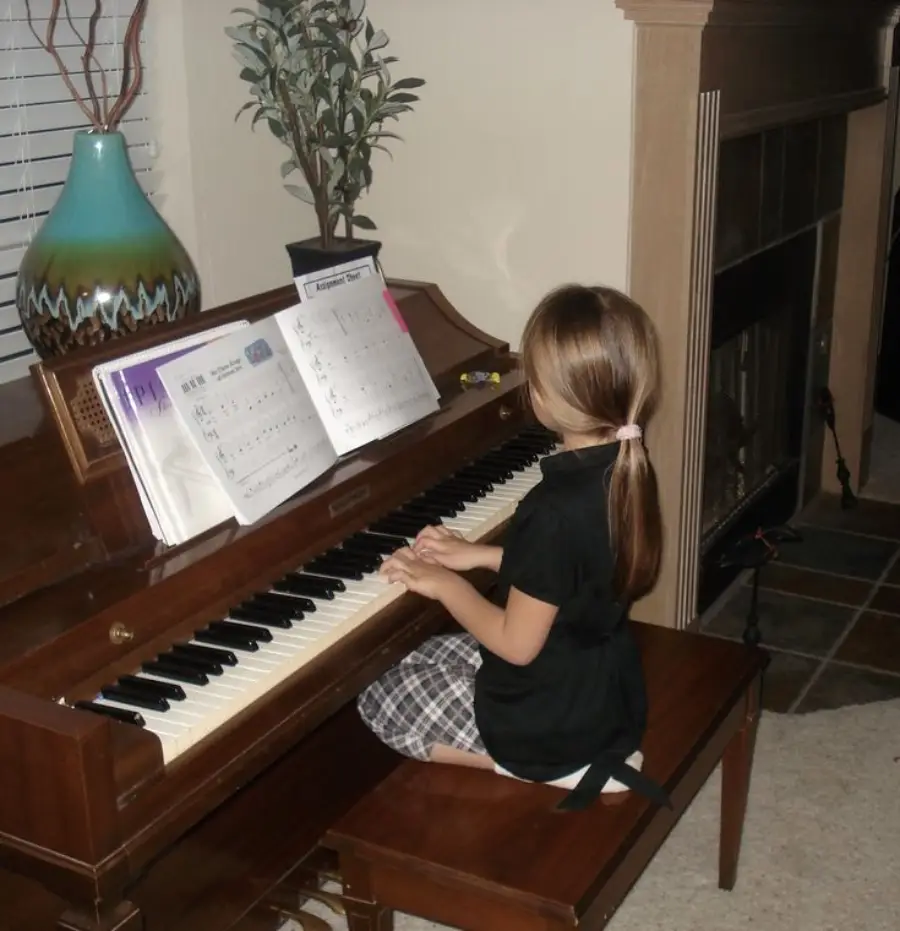 A young girl with brown hair in a ponytail, wearing a black shirt and plaid skirt, sits at a piano playing music. Sheet music is open in front of her, and a large vase is on top of the piano.