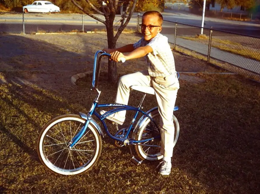 A boy wearing glasses and light-colored clothes smiles while posing on a blue bicycle in a grassy yard with a tree and a street in the background.