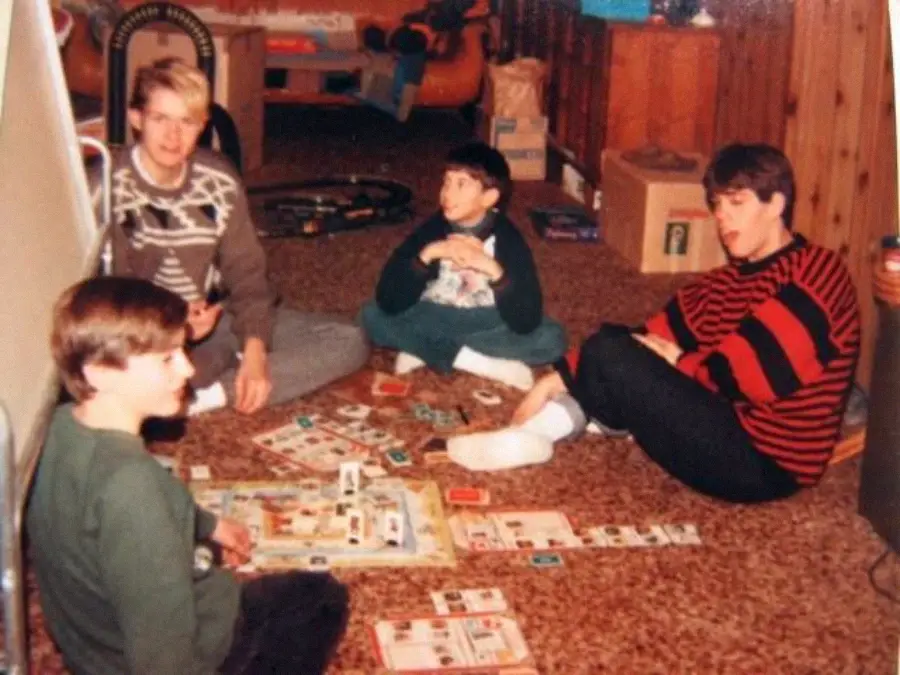 Four people sit on a carpeted floor playing a board game. Game cards, money, and pieces are spread out between them. They are in a wood-paneled room with boxes and toys in the background.