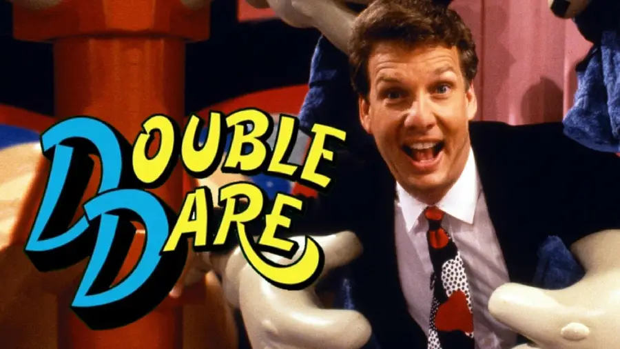 A smiling man in a suit stands next to the colorful "Double Dare" TV show logo, with cartoon-like set decorations in the background.