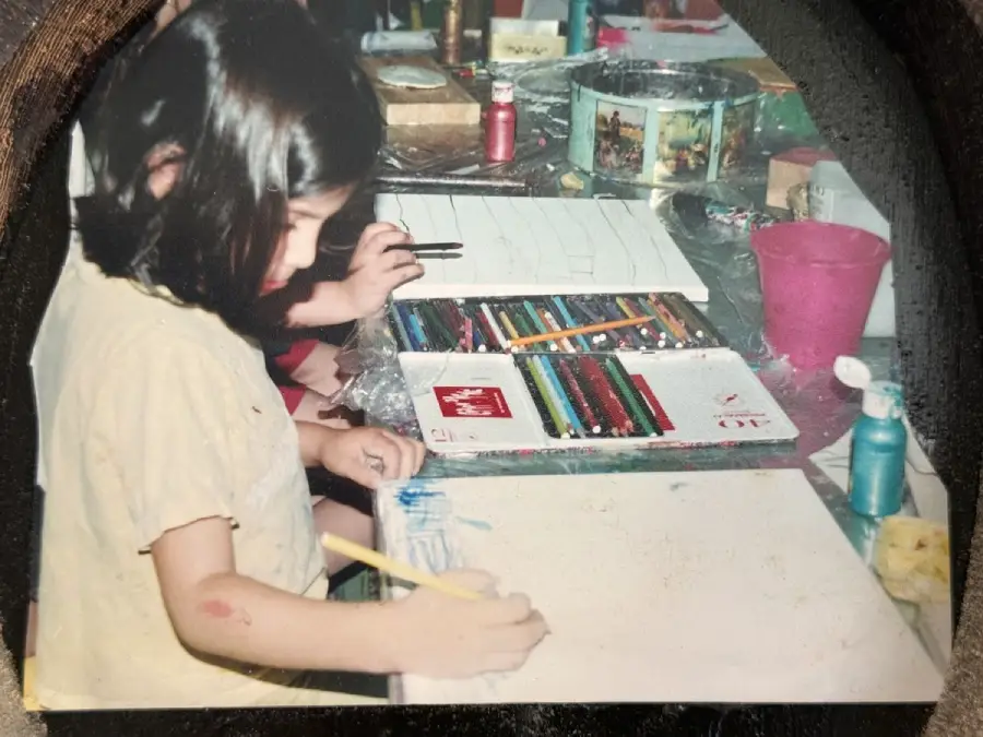 A child with shoulder-length hair draws on a piece of paper at a table covered with colored pencils, paints, and art supplies. Other art materials and a pink cup are visible in the background.