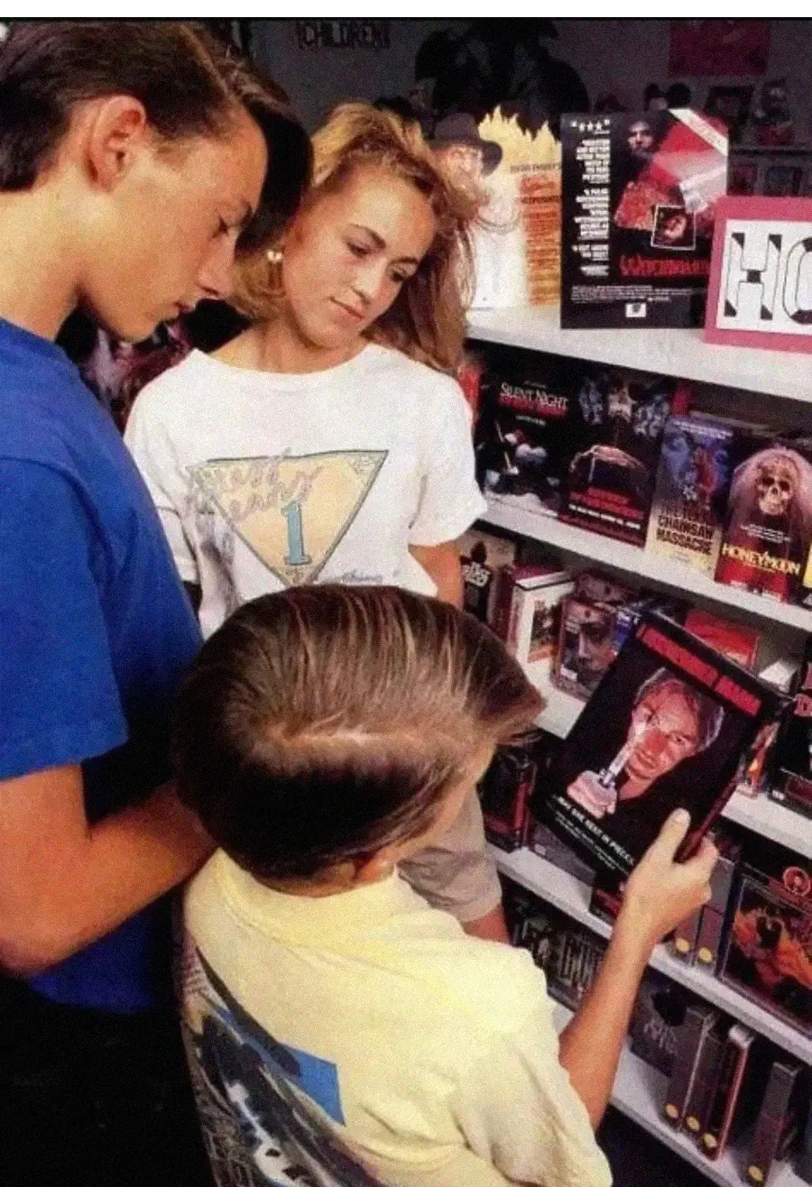 Three kids stand in a video rental store looking at horror VHS tapes; one holds up a tape with a scary face on the cover, while the others watch. Various horror movie boxes are visible on the shelves.