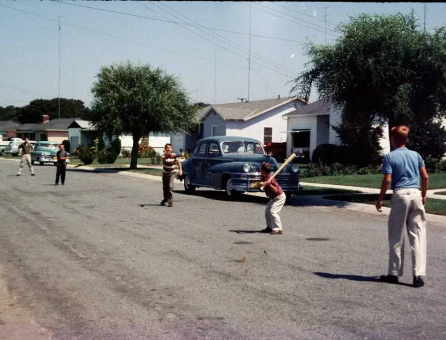 Children play a game of baseball in the middle of a suburban street lined with houses and vintage cars on a sunny day.