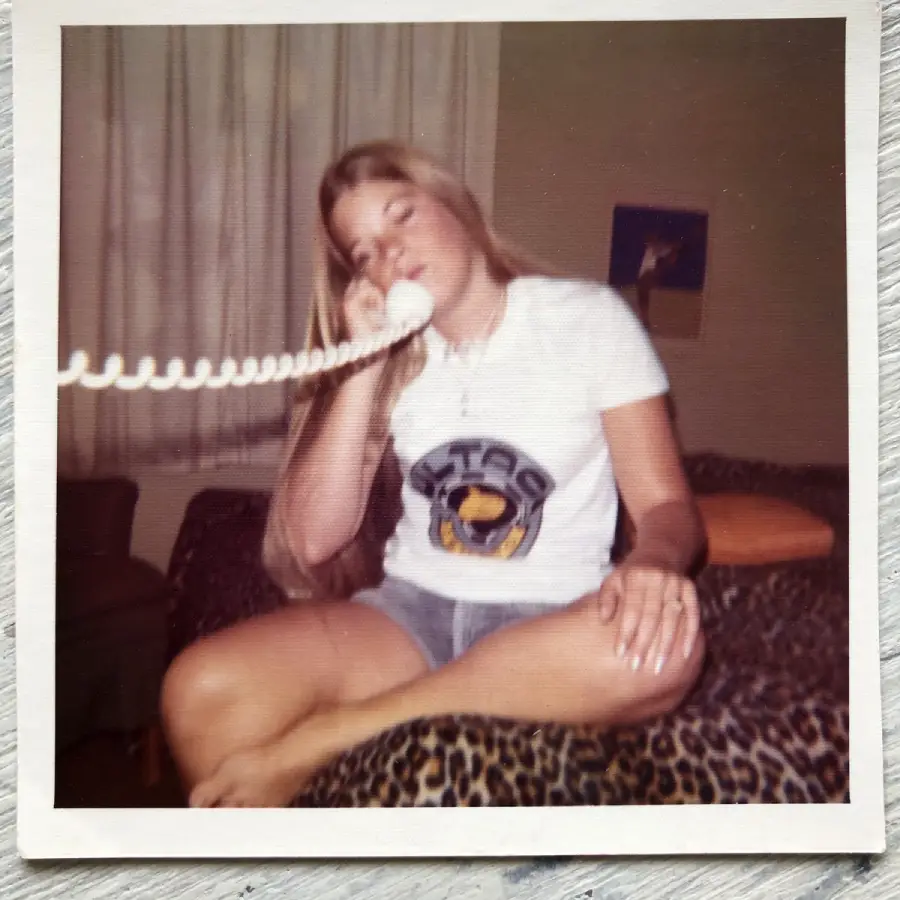 A young woman with long blonde hair sits cross-legged on a bed with a leopard print blanket, talking on a corded phone. She wears a white t-shirt and shorts, with a relaxed and casual expression.
