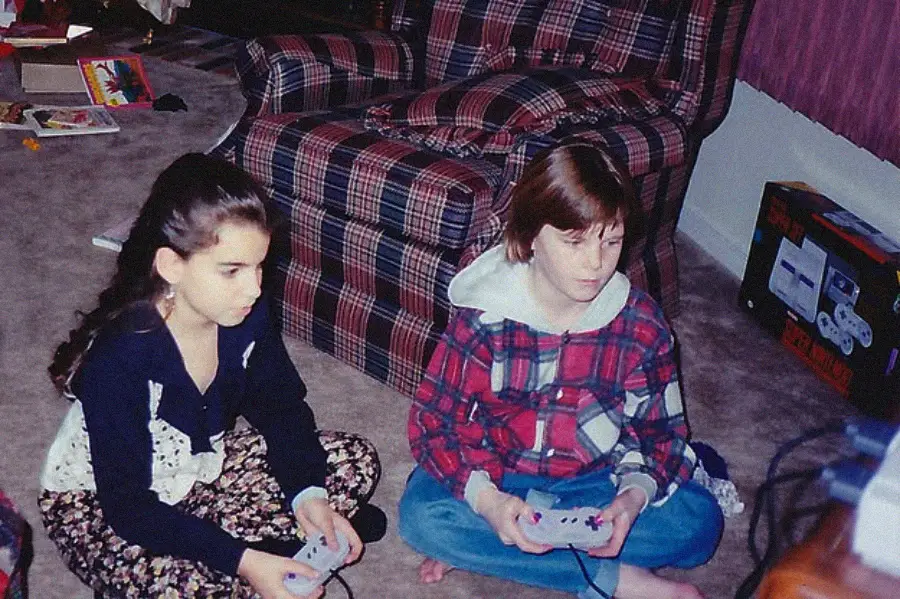 Two children sit on the carpeted floor playing a video game with controllers. Behind them is a plaid armchair and a Super Nintendo console box. The room has scattered toys and a cozy, lived-in feel.